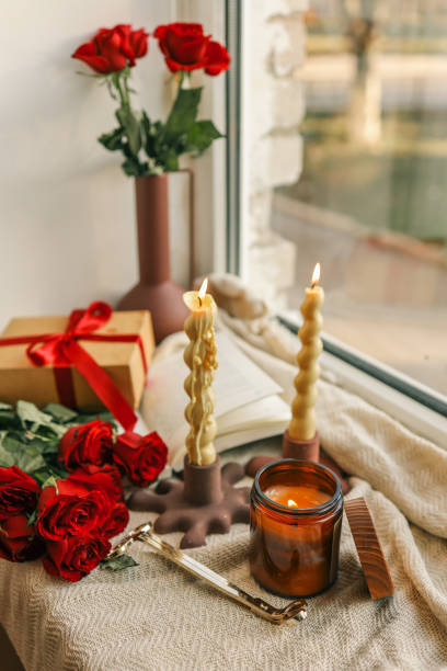 istock Two twisted candles burning in modern holders by a windowsill, accompanied by a red ribbon gift box, red roses, and a vase with roses, creating a cozy ambiance with natural light 2194324849