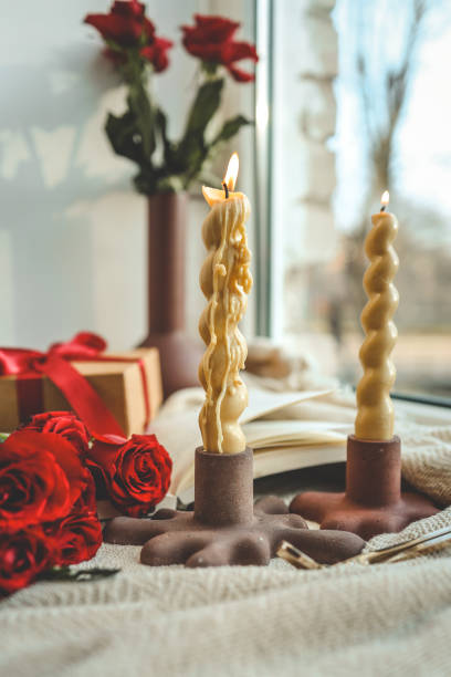 Two twisted candles burning in modern holders by a windowsill, accompanied by a red ribbon gift box, red roses, and a vase with roses, creating a cozy ambiance with natural light