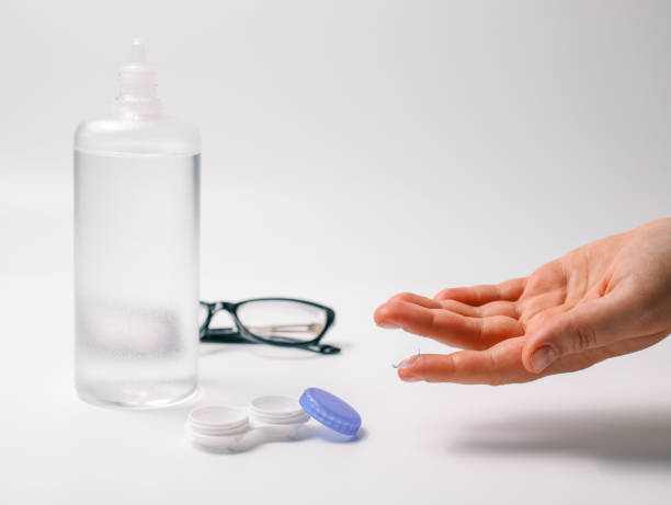 istock Hand holding a transparent contact lens next to lens case, solution bottle, and glasses on a white background. Concept of eye care, vision correction, and optical hygiene 2194325477