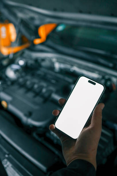 istock Mechanic holding a smartphone with a blank white screen in front of an open car engine for diagnostics or maintenance. Indoor workshop setting 2194327363