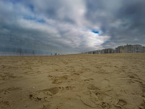 A scenic beach landscape featuring a reflective water body, adjacent modern buildings, and dramatic overcast clouds. Captures the tranquility and serenity of coastal living with a touch of dramatic weather.