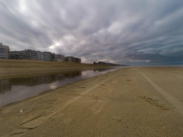 A scenic beach landscape featuring a reflective water body, adjacent modern buildings, and dramatic overcast clouds. Captures the tranquility and serenity of coastal living with a touch of dramatic weather.