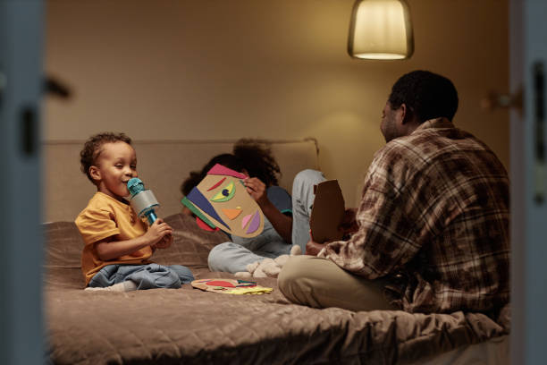 Shot through open bedroom doors of funny little Black boy singing into small microphone having fun over karaoke with dad and sister, while sitting together on bed