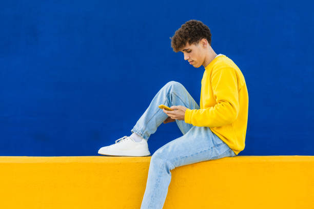 istock Young man using a smartphone sitting on a yellow wall with blue background 2194355179