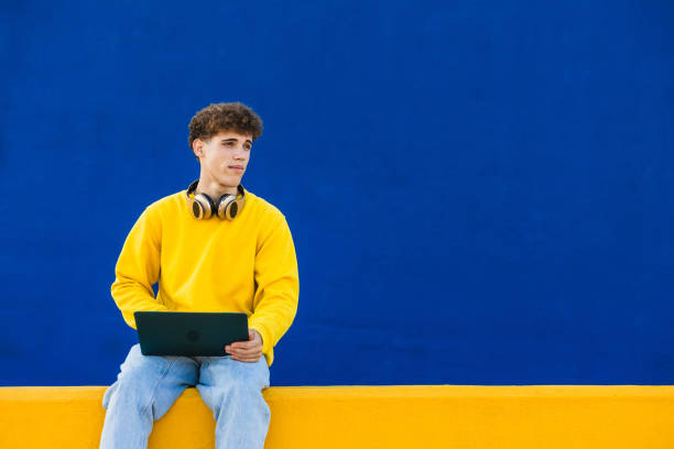 istock Young man using laptop with headphones around neck in front of blue wall 2194355195