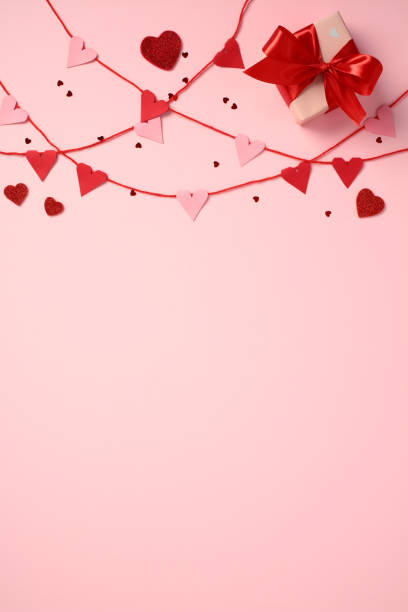 Valentine's Day vertical background with red and pink paper hearts on strings, scattered heart confetti, and a gift box with a red ribbon on a pink background