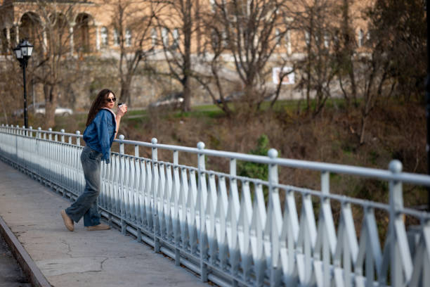 Beautiful young woman is having coffee in a white cup outdoors on a bridge with a city view background