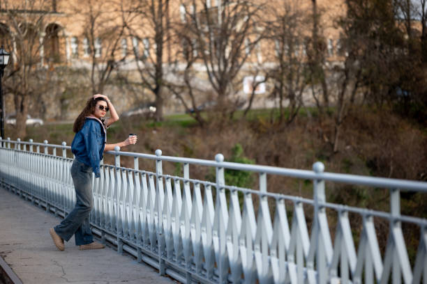 Beautiful young woman is having coffee in a white cup outdoors on a bridge with a city view background