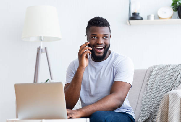 istock Man Smiling While Talking on Phone and Working on Laptop at Home Office in Bright Room 2194366749