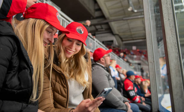 Two young women share a laugh while looking at a phone during an exciting ice hockey match
