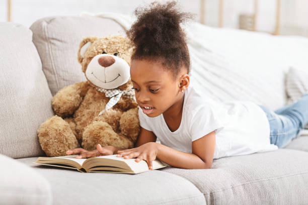 istock Young Child Reading a Book While Relaxing With a Teddy Bear in a Cozy Living Room 2194369379