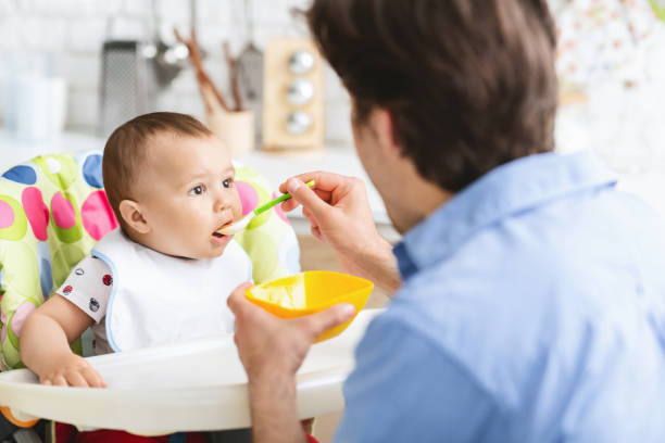 istock Father Feeding Baby in High Chair During Mealtime in a Bright Kitchen Setting 2194370325