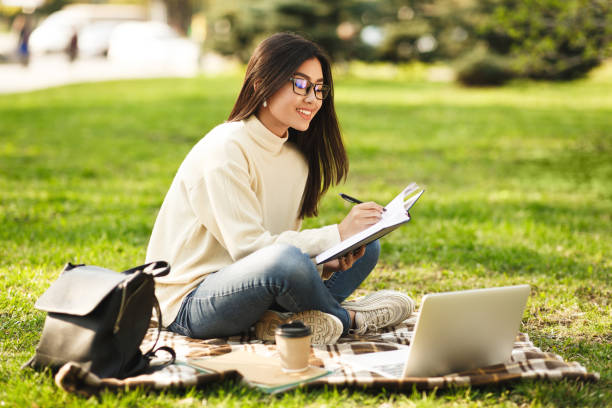 istock Young Woman Sitting on Grass Writing in Notebook While Using Laptop Outdoors 2194372162