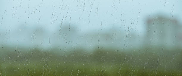 istock Atmospheric minimal backdrop with rain droplets on glass. Wet window with rainy drops and dirt spots closeup. Blurry buildings and green trees under blue sky against dirty window glass with raindrops. 2194374677