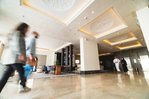 Spacious hotel lobby with elegant decor, featuring blurred movements of diverse guests. Beige marble floors and intricate ceiling designs create a luxurious ambiance. Guests are dressed in casual and formal outfits, walking across the well-lit, contemporary space.