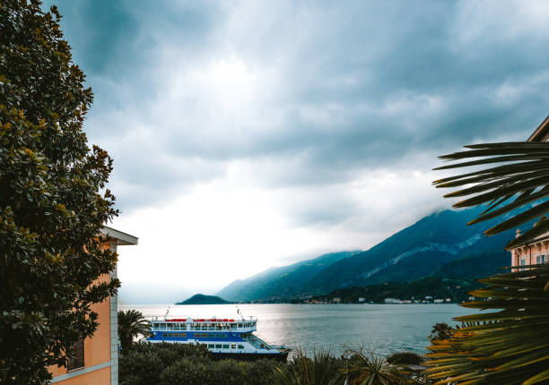 Ferry boat docking at the pier in Bellagio, a charming village on Lake Como, under a dramatic cloudy sky, with the Grigne mountains in the background, Lombardy, Italy