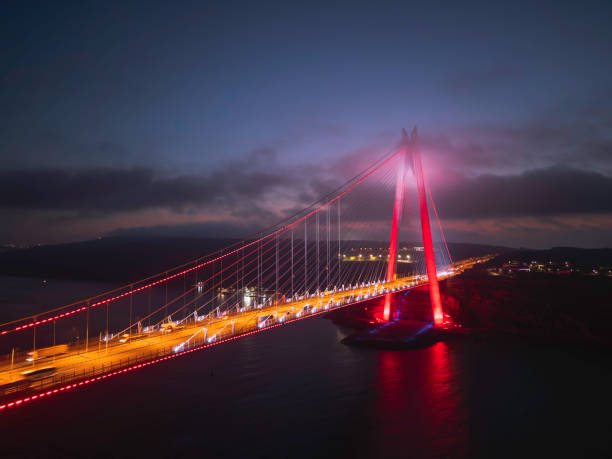 A stunning nighttime view of the Yavuz Sultan Selim Bridge, also known as the Third Bosphorus Bridge, spanning the Bosphorus Strait in Istanbul, Turkey. The modern cable-stayed bridge is illuminated in red and white lights, reflecting beautifully on the dark waters below. This architectural marvel connects Europe and Asia, serving as a major transportation route while standing as a symbol of Istanbul’s blend of tradition and modernity.
