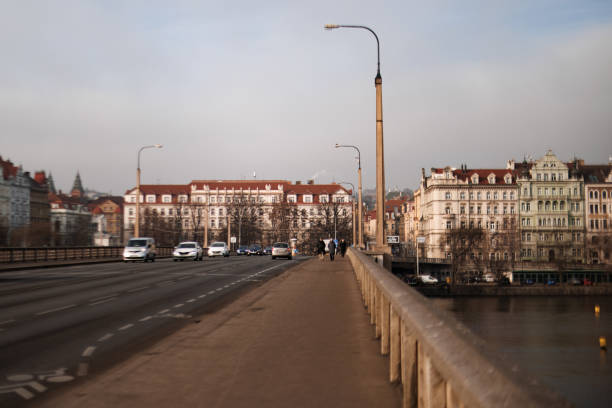 Tourists walking across Jirasek Bridge in Prague, Czech Republic, with cars passing by on a cloudy day. The bridge spans the Vltava River and connects Smíchov with the city center.