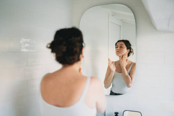 istock Woman applying moisturizer on face looking in the mirror in bathroom 2194400405
