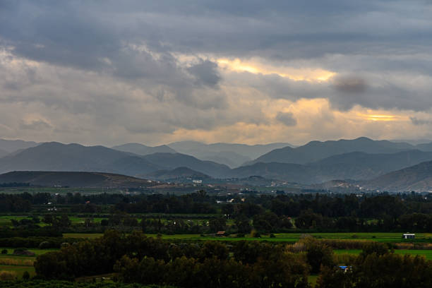 Golden light breaks through clouds, illuminating rolling hills and lush fields in a tranquil countryside setting.