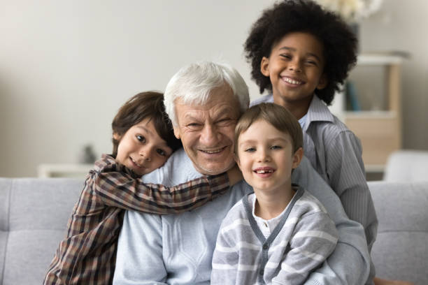 Three multiracial grandsons hug their hoary great-grandfather, smile look at camera, sitting on sofa posing together for family photo. Multigenerational family bond, ties, unity. Offspring portrait