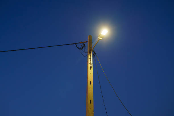 Illuminated streetlamp mounted on a wooden pole against a clear blue sky.