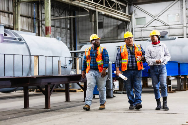A multiracial group of three workers conversing as they walk through a steel fabrication factory. The mature Hispanic man in the middle, is talking and holding a rolled up set of blueprints. The two young workers walking beside him are listening and looking toward him. One is an African-American woman.