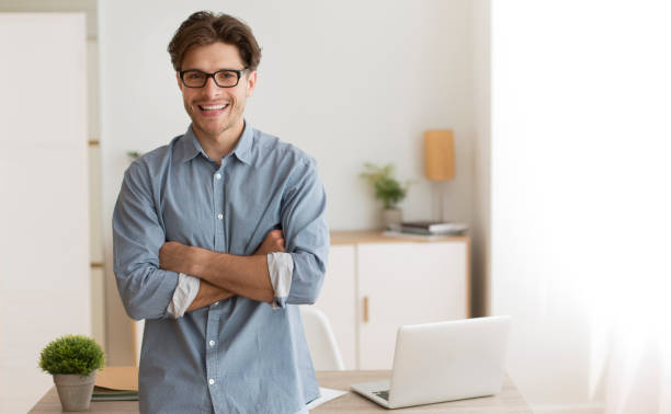 Man Smiling in a Bright Office While Standing Confidently by a Desk With a Laptop