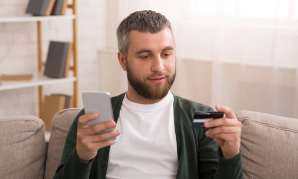 istock Young Man Shopping Online Using Smartphone and Credit Card While Sitting on Couch 2194458538