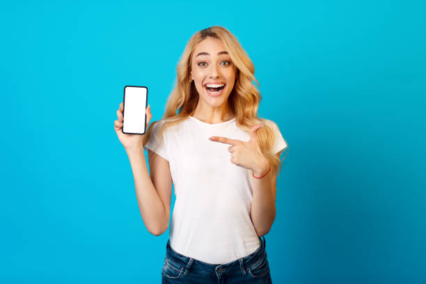 istock Young Woman Smiles and Points at Smartphone Against a Blue Background in a Bright Setting 2194461146