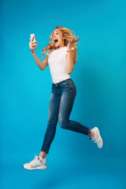 Young Woman Jumping With Excitement While Holding a Smartphone Against a Bright Blue Background