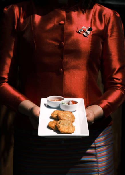 This image showcases the traditional Thai fish cakes, Tod Mun Pla, served on a white rectangular plate accompanied by two dipping sauces, offering a delightful combination of flavors. The person holding the dish is dressed in vibrant traditional Thai clothing, adding an authentic cultural touch. The composition highlights the intricacies of Thai cuisine and presentation, making it an ideal depiction of a culinary experience in a Thai restaurant.
