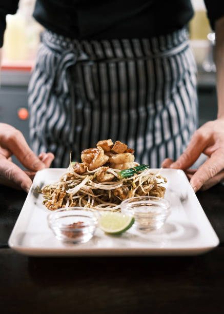 This image showcases a classic serving of Thai Pad Thai noodles, complete with stir-fried shrimp, fresh bean sprouts, peanuts, and garnished with lime. The dish is accompanied by small bowls of condiments, allowing for customized flavors. Served on a pristine white plate, the meal is carefully presented by a chef in a striped apron, emphasizing the artistry and authenticity of Thai cuisine. The composition highlights the vibrant colors and textures, perfect for a cozy and flavorful dining experience in a traditional Thai restaurant.