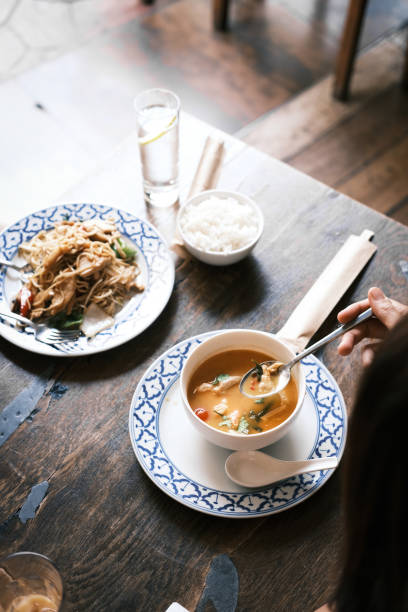 This image captures a beautifully arranged Thai meal featuring red curry with prawns and stir-fried egg noodles with beef. The red curry, served in a white bowl with a vibrant broth and fresh herbs, pairs perfectly with the stir-fried noodles garnished with vegetables and Thai spices. The dishes are presented on matching blue-patterned plates, accompanied by steamed rice, reflecting the authenticity and elegance of Thai cuisine. The wooden table and natural lighting create a warm, inviting restaurant ambiance, perfect for enjoying these traditional Thai flavors.