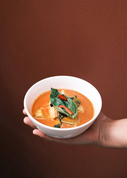 This image captures the essence of traditional Thai red curry, presented in a white bowl held by hand. The curry is rich in color, showcasing its creamy coconut base, bold spices, and a medley of fresh vegetables, topped with fragrant basil leaves. The warm brown background enhances the vibrant orange hues of the dish, making it visually appealing. Perfectly styled, this dish reflects the authentic flavors of Thai cuisine, highlighting its balance of spiciness, sweetness, and aroma, making it ideal for lovers of gourmet Asian food.