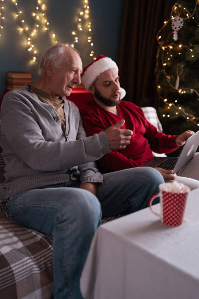 istock Bearded man in Santa hat teaches elderly father how to use laptop and internet while sitting on couch at home on Christmas 2194484779