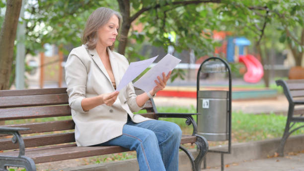 istock Old Woman Reading Documents while Sitting Outdoor on Bench 2194485690