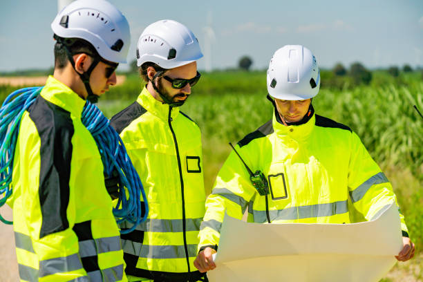 Team of engineers examining plans while dressed in safety gear under a clear sky in a rural area