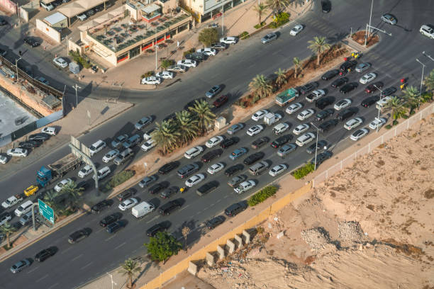 High angle view of dense traffic in Riyadh, featuring a mix of cars on a wide road bordered by palm trees. Buildings and sandy terrain complete the sunny urban scene.