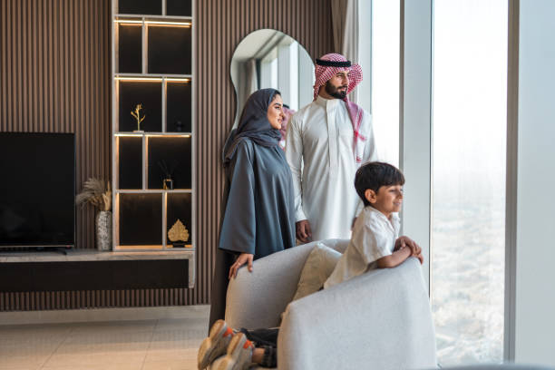 A diverse Arabic family enjoys a majestic view of Riyadh from their contemporary home. The father, mother, and young son display happiness and unity while gazing through large windows in a stylish living room.
