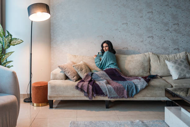 Young Arabic female enjoying leisure time on a plush sofa in a contemporary home, surrounded by soft blankets and a warm floor lamp, creating a serene atmosphere for relaxation and comfort.
