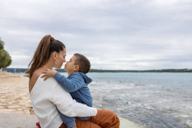 A loving moment shared between a mother and her child while sitting by the ocean on a cloudy day, showcasing parental affection and the serene coastal environment.
