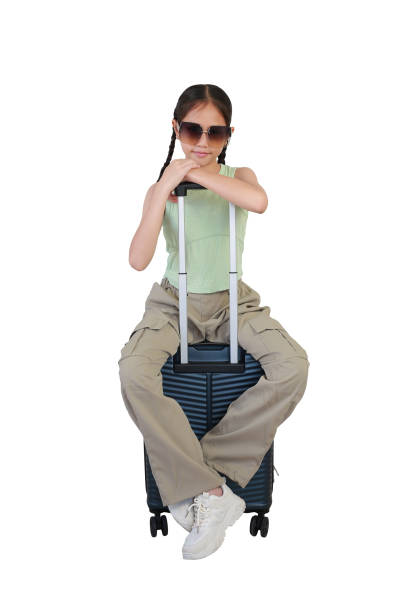 Asian girl child sitting on suitcase posing and looking straight camera while wearing sunglasses isolated on white background. kid fashion and travel concept. Full body photo.