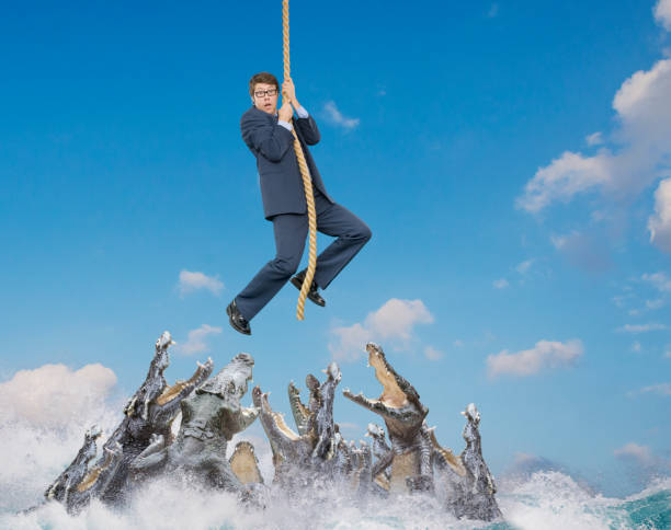 Businessman hangs on end of rope above snapping crocodiles, John M.Lund