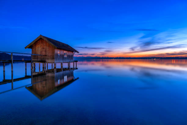 A rustic wooden and tin boat shed stands at the end of a long lakeside walkway on Lake Starnberg in southern Germany. An old tin dinghy sits inside, surrounded by clear, shallow water that reflects the natural scenery. Forested hills rise in the distance, creating a calm and picturesque outdoor setting.
