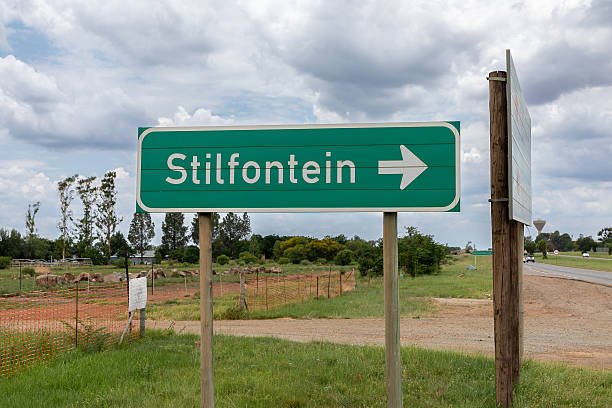 Green Road sign reading Stilfontein with a white arrow pointing to the right.  Stilfontein is a town in South Africa.  \nSign against a cloudy sky.