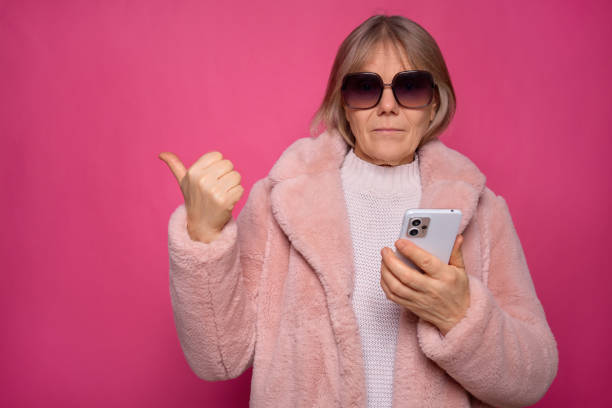 istock A woman in a stylish pink coat and oversized sunglasses shows a thumbs up while using her smartphone. The lively pink background adds a cheerful touch to the scene 2194539729