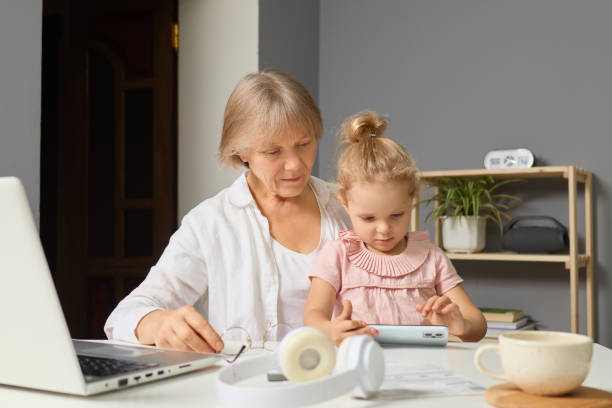 istock A grandmother and her young granddaughter sit at a table, engaged in a shared activity. The girl interacts with a device while her grandmother observes with joy, creating a warm family moment 2194545722