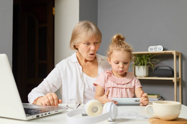 A grandmother and her granddaughter sit together at a table, focused on a digital device while surrounded by a laptop and a cup. This intimate moment captures their shared enjoyment and curiosity