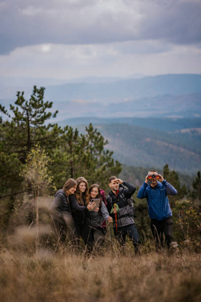 A group of five friends stands on a mountain trail, using a tablet to navigate their way. The cloudy sky looms overhead, and the landscape stretches into the distance.